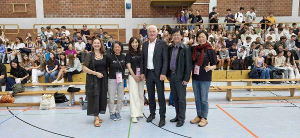 Knoten der Kulturen Beteiligte Personen vor einer mit Schülerinnen und Schülern besetzten Tribüne in der Turnhalle