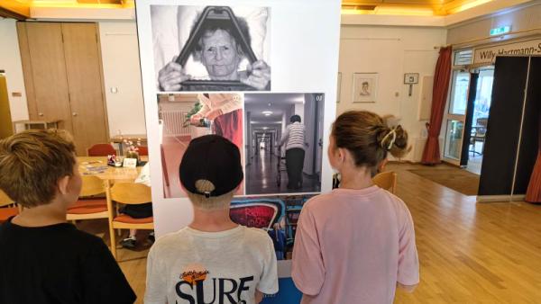 3 Kinder betrachten ein Bild einer Seniorin an einer Ausstellungswand