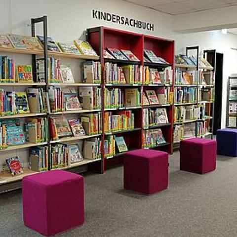 Eine lange Bücherwand mit Kinderbüchern, davor stehen 4 quadratische Stoffhocker ©Stadtbibliothek Kempen